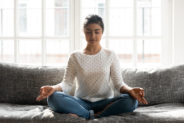 Foto achtsame junge indische frau in voller länge, die eine mudra-geste macht, die zu hause in lotusposition auf einer bequemen couch sitzt friedliches tausendjähriges mädchen, das tief meditiert und allein atem-yoga-übungen macht