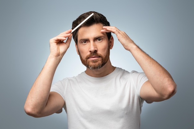 Acicalamiento perfecto guapo europeo barbudo hombre de mediana edad cepillando el cabello con peine y mirando