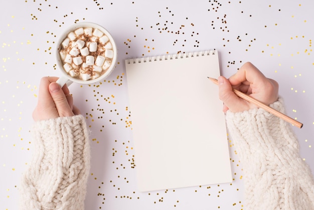 Acima em cima, close-up, vista da foto de mãos femininas usando uma caneta de madeira, escrevendo um texto para o caderno de desenho em branco aberto, bebendo uma bebida saborosa