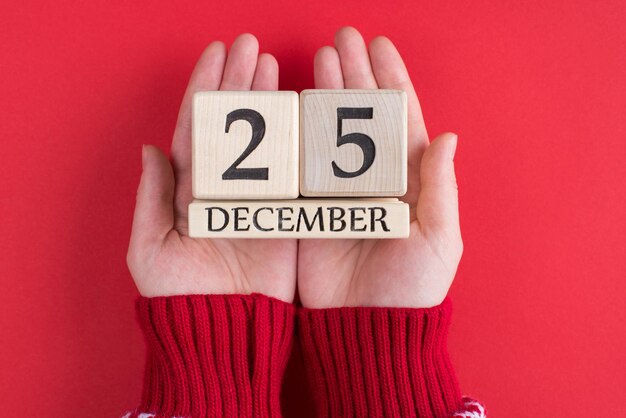 Acima em cima da cabeça, feche a foto da visão em primeira pessoa de uma adolescente com mãos segurando um calendário de madeira com a data de natal isolada sobre fundo vermelho