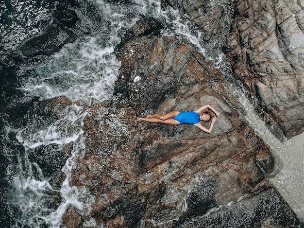 Acima, foto de uma bela jovem em um maiô azul deitado sobre a rocha na maré alta. Conceito de férias de verão