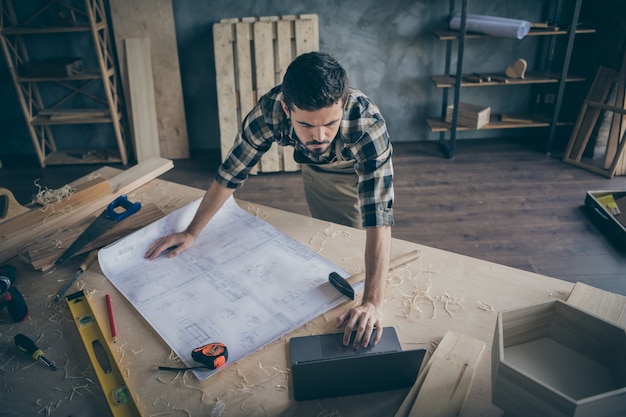 Acima, o trabalhador de madeira focalizado de alto ângulo tem uma tela com um plano de construção civil, use o computador assistir a vídeos de trabalhos manuais na garagem de casa