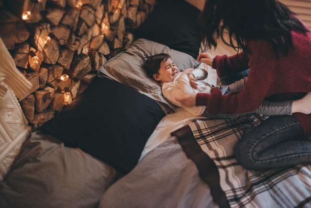 Acima tiro de mãe feliz e sua filha brincando na cama com decoração aconchegante e luzes de natal mulher bonita e menina bonitinha se divertem no fim de semana de inverno pessoas da família natal