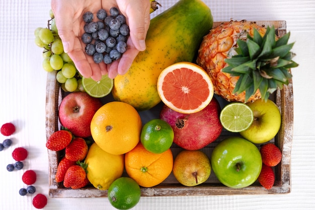 Acima vista da cesta de madeira cheia de frutas frescas coloridas. Mãos de mulher madura segurando um grupo de mirtilos. Alimentação e estilo de vida saudáveis. fundo branco