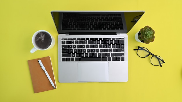 Acima vista do computador portátil, xícara de café, óculos e notebook em fundo amarelo.