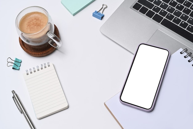 Acima vista smart phone, xícara de café, laptop e bloco de notas na mesa de escritório branca.