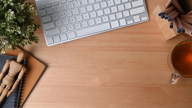 Acima vista teclado sem fio, xícara de chá, porta-lápis e caderno na mesa de madeira.