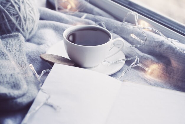 Acogedor bodegón de invierno: taza de té caliente y libro con cuadros calientes en el alféizar de la ventana