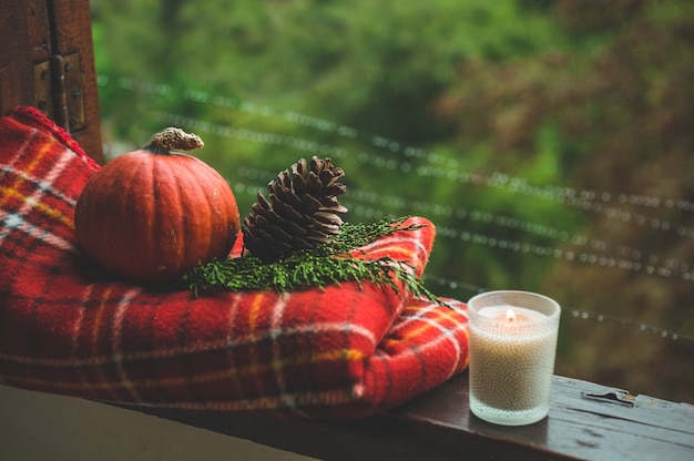 Acogedor bodegón de otoño: taza de café caliente y libro abierto en el alféizar de la ventana vintage con manta roja, calabaza, piña, velas y lluvia afuera. Otoño. Departamento. Lluvia