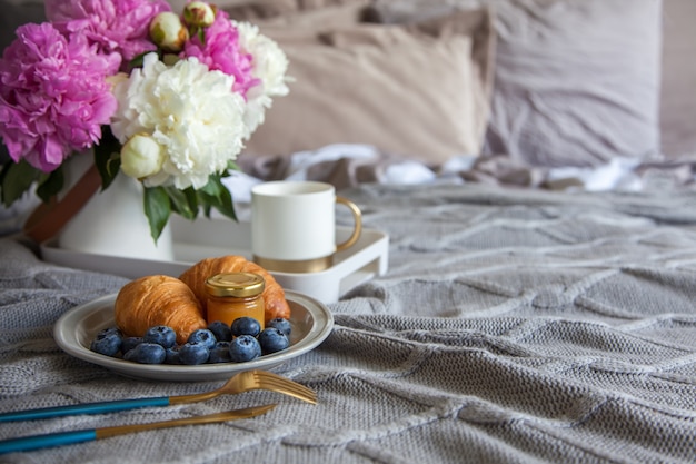 Acogedor desayuno en la cama, taza de café, arándanos, mermelada y croissants en la cama gris. Piones rosados y blancos en el jarrón.