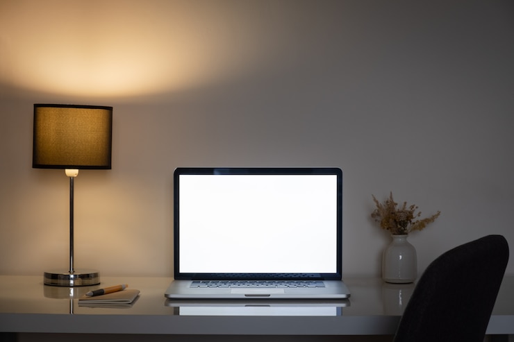 Foto acogedor escritorio de oficina en casa por la noche iluminado por una lámpara y una pantalla de portátil blanco.