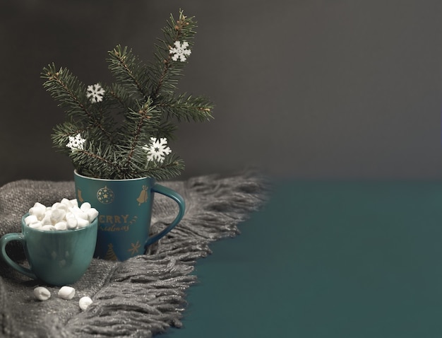 Acogedor hogar hugge Fondo de Navidad o año nuevo con taza de Navidad con ramas de abeto, copos de nieve, taza de café o cacao con malvaviscos en cuadros de punto gris en la oscuridad. Enfoque selectivo. Copie el espacio.