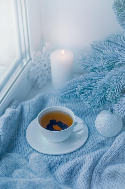 Acogedor invierno bodegón taza de té caliente y libro con cálido plaid en el alféizar de la ventana contra el paisaje nevado desde el exterior