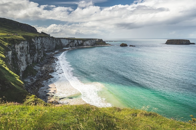 Acogedora bahía junto a la costa de Irlanda del Norte, el acantilado cubierto de hierba bañada por el mar turquesa