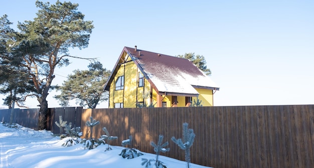 Una acogedora casa amarilla en la nieve en invierno en el pueblo está rodeada de pinos Techo cubierto de nieve, calefacción y ventilación, ventanas trapezoidales