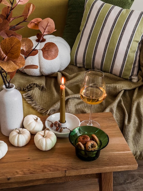 Acogedora casa bodegón Una copa de vino, galletas, decoración otoñal, vela encendida sobre una mesa de madera cerca del sofá con almohadas y mantas