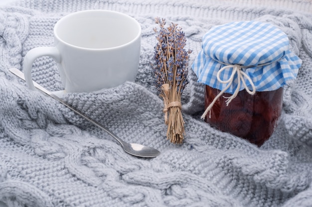 Acogedora casa, hora del té, taza blanca con cuchara, rama de lavanda y mermelada en un frasco de cuadros tejidos.