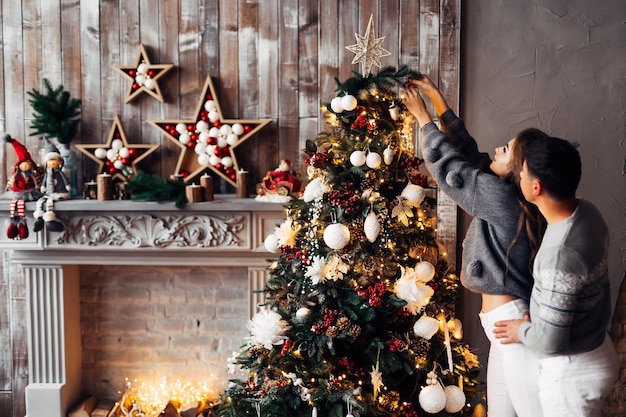 Acogedora habitación con chimenea decorada y árbol de Navidad el chico abraza a la chica por la cintura y ella decora el árbol de Navidad