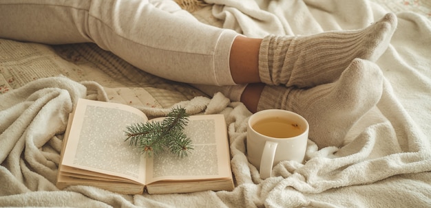 Acogedora noche de invierno, cálidos calcetines de lana. La mujer está acostada pies sobre una manta lanuda blanca y un libro de lectura. Escena de ocio acogedor. El texto del libro no se puede leer. Mujer relajante en casa. Estilo de vida cómodo.