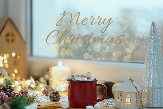 Acogedora tarjeta de navidad casera con texto feliz navidad vista de ventana taza de chocolate caliente en el alféizar de la ventana
