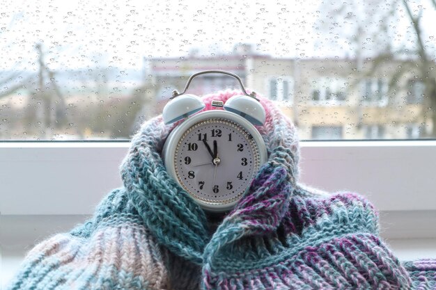 Foto acogedores días de invierno, un reloj de alarma blanco envuelto en un artículo de punto se encuentra en un espacio de primer plano del alféizar de la ventana para el texto