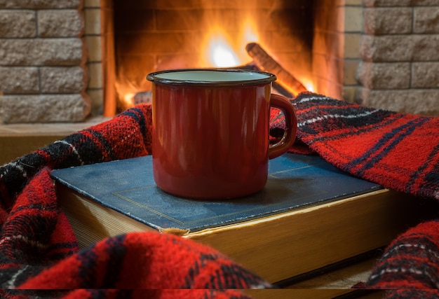 Aconchegante cena antes da lareira com caneca esmaltada vermelha com chá, um livro e cachecol de lã.