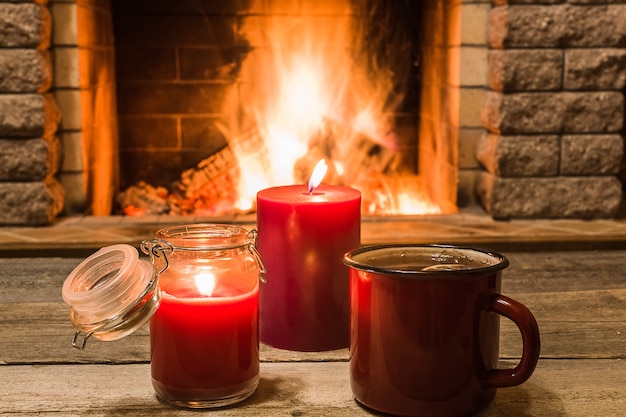 Aconchegante cena contra lareira com caneca esmaltada vermelha com chá e duas velas.