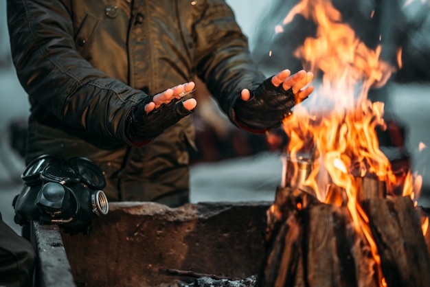 Acosador, persona del sexo masculino calienta sus manos en el fuego. Estilo de vida postapocalíptico con máscara de gas, apocalipsis,