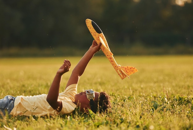 Acostado con un avión de juguete en el césped Un niño afroamericano se divierte en el campo durante el día de verano