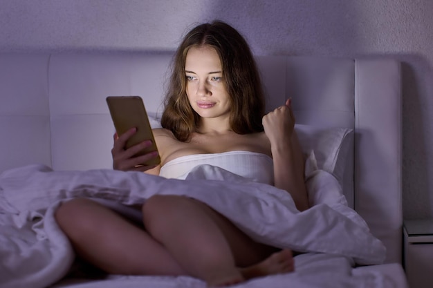 Acostado en la cama, la mujer mira la pantalla del teléfono inteligente en la habitación oscura, la luz del teléfono ilumina el rostro femenino