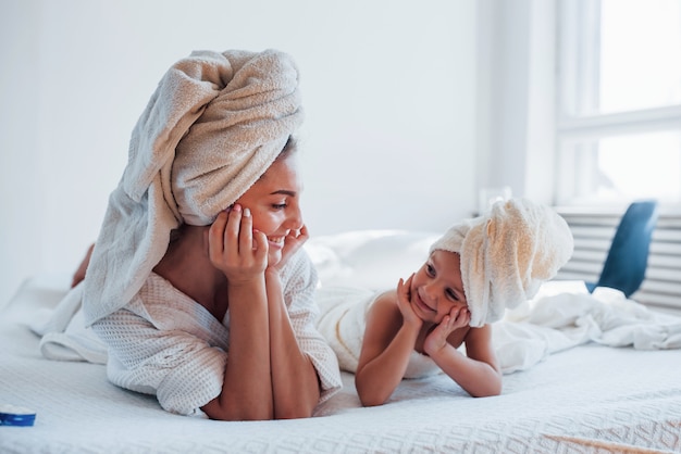 Acostado juntos en la cama blanca. La joven madre con su hija tiene un día de belleza en el interior de la habitación.