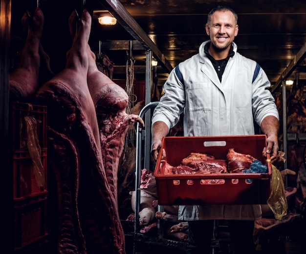 Foto açougueiro alegre em roupas de trabalho segurando uma caixa com pedaços de carne em pé no meio de carcaças de carne