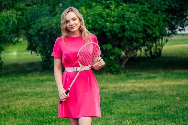 Activa alegre hermosa joven rubia en un estilo vestido rosa jugando raqueta de bádminton de tenis en el parque de verano. picnic y estilo de vida activo en concepto de juventud