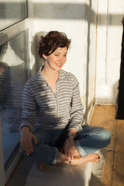 activa, hermosa mujer de mediana edad con una hermosa sonrisa. Sentado en el alféizar de la ventana cerca de la ventana. Reflexión en la ventana.