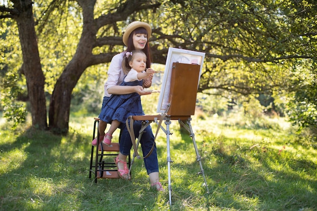 Actividad al aire libre para niños, hobby y concepto de creatividad. Madre atractiva joven enseña a su pequeña hija a pintar sobre caballete en el parque o jardín soleado de verano.