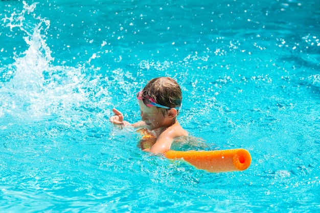 Actividad de verano al aire libre Concepto de vacaciones de salud divertidas Niño sonriente feliz de cinco años en gafas de natación se ríe y se cierra de salpicar agua en la piscina con fideos en un caluroso día de verano