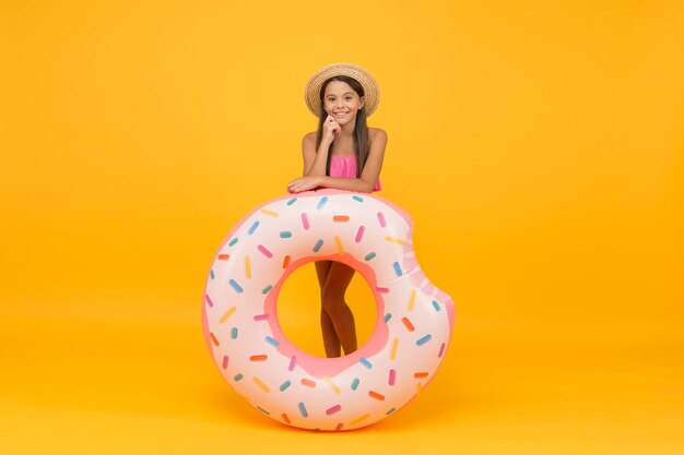 Actividad de verano en la playa. infancia feliz. niña pequeña usa sombrero de paja y traje de baño. niño sonriente sobre fondo amarillo. vacaciones y vacaciones. fiesta de piscina. niño despreocupado con anillo de goma.