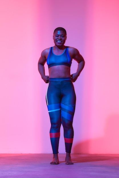 Foto actividade física de corpo inteiro retrato vertical positivo jovem mulher negra nos esportes top sutiã e perneiras posando no estúdio olhando para a câmera isolado em rosa néon fundo do estúdio banner