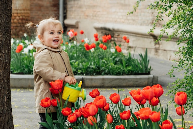 Actividades de jardinería de primavera para niños niña pequeña linda en impermeable regando tulipanes rojos