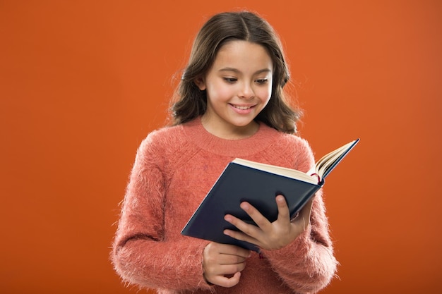 Actividades de lectura para niños. Chica sostenga el libro leyó la historia sobre fondo naranja. El niño disfruta leyendo el libro. Concepto de librería. Maravillosos libros para niños gratuitos disponibles para leer. Literatura infantil.