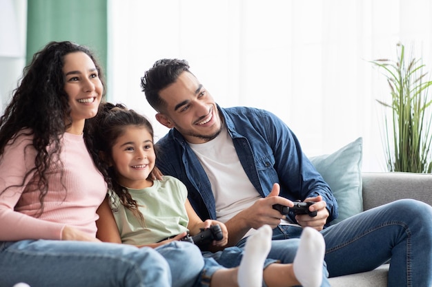 Actividades de tiempo en familia. Feliz madre y padre árabes jugando videojuegos con su pequeña hija mientras se relaja en el sofá en casa, papá y linda chica sosteniendo joysticks, compitiendo entre sí, espacio libre