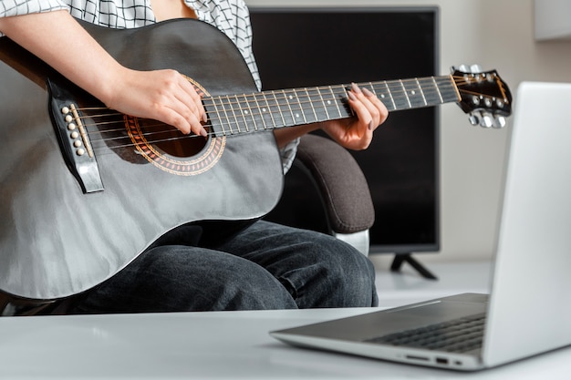 Actuación de guitarra musical online. Mujer joven toca la guitarra acústica en casa para la audiencia en línea en la computadora portátil. Video concierto o curso de clases en línea entrenamiento de guitarra música e educación durante el encierro.