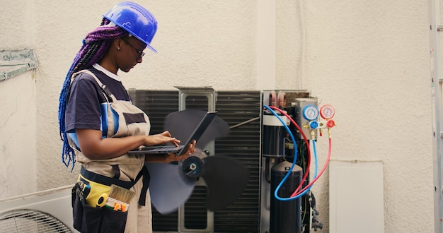 Adepto reparador afroamericano trabajando en aire acondicionado exterior con portátil. Trabajador competente que optimiza el rendimiento del sistema HVAC y garantiza que funcione a su máxima capacidad