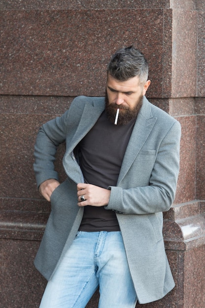 adicción al tabaco. carismático hombre de negocios fumando al aire libre. hombre barbudo fuma cigarrillo. brutal hipster maduro con chaqueta. hombre con vello facial arreglado. chico con peinado elegante. Estilo de otoño.