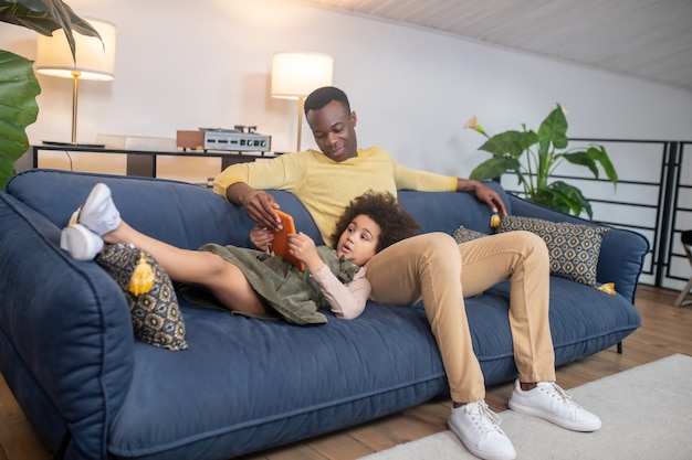 Foto adicción a internet. hombre de piel oscura adulto joven sentado recogiendo tableta de indignada hijita acostada en el sofá en casa