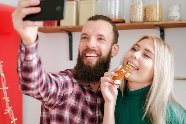 Adicción a las redes sociales. Pareja divertida con smartphone para tomar selfie con hombre de pan de jengibre en la cocina.