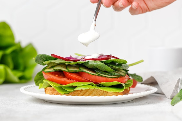 Adicionando creme de molho no sanduíche vegano de torrada vegetal com tomate espinafre pepino Sanduíche