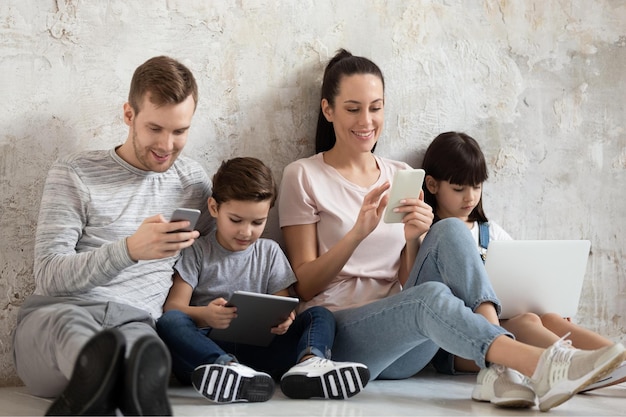 Adicto a la tecnología feliz padre milenario madre y dos hijos hermanos sentados en un cálido suelo de madera usando teléfonos móviles, computadora y tableta digital relajándose juntos en casa