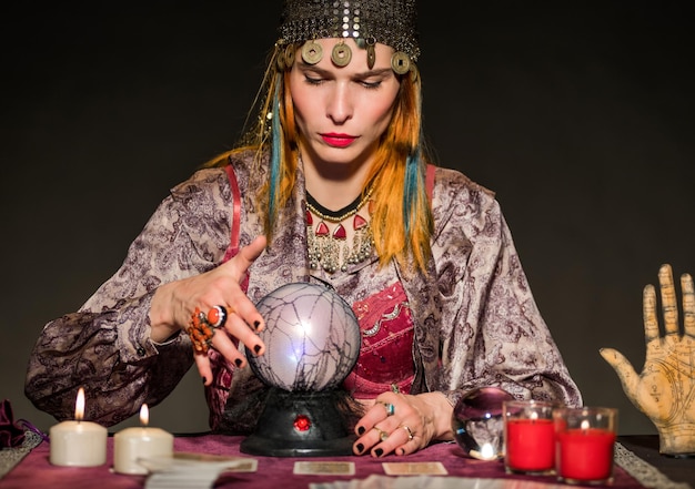 Adivino serio en traje auténtico sentado en la mesa con velas encendidas y mirando la bola mágica mientras predice el futuro en fondo negro