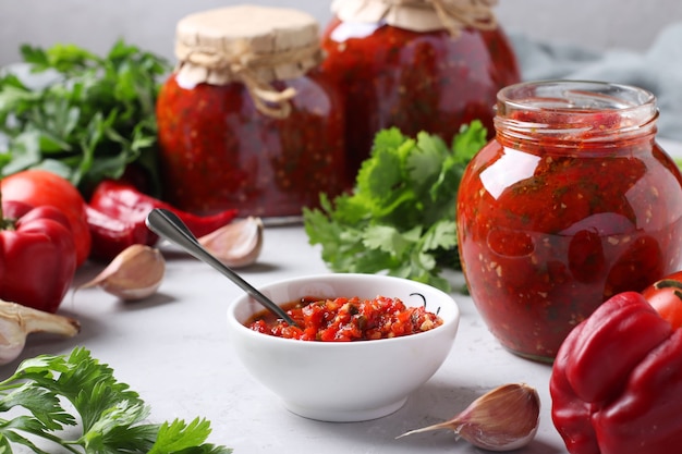 Adjika armênio de pimenta vermelha, tomate, alho, coentro e salsa em uma tigela e potes, bem como ingredientes frescos em uma mesa. Ainda vida na superfície cinzenta.